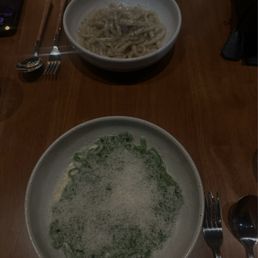 Cacio E Pepe - Two bowls of pasta, one with creamy noodles and the other with a green vegetable dish, are placed on a wooden table in a restaurant setting.