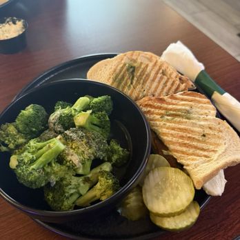 McBride's Morris - Panini with side of broccoli - A meal is served on a dark plate with two grilled sandwiches, featuring grill marks and green herbs. A bowl of steamed broccoli and sliced pickles are also on the plate. The setting appears to be a restaurant with a wooden table, and additional dishes are visible in the background. - American Near Me - Morris, Illinois