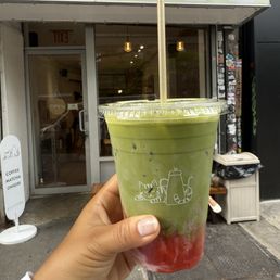 Iced strawberry matcha latte - A hand is holding a plastic cup containing a layered iced matcha latte with green and red sections, topped with a straw. The background shows the entrance of Kijitora café, with a glass door, an 'OPEN' sign, and a small outdoor sign listing coffee, matcha, and onigiri. The café has a modern exterior with a light-colored awning and some visible interior lighting.