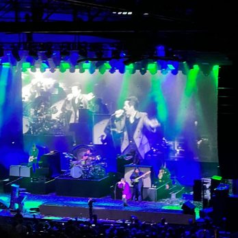 St Augustine Amphitheatre - A live music concert at an indoor venue features a band performing on stage with multiple musicians, including a singer, guitarist, bassist, and drummer. The stage is lit with green and blue lighting, and a large screen behind the performers displays a black-and-white image of a singer holding a microphone. Audience members are visible in the foreground, and sound equipment is set up around the stage. - Music Venues Near Me - Saint Augustine, Florida