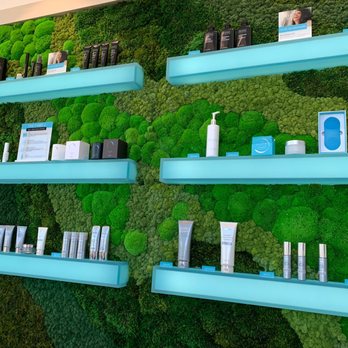OVME - Wall of products - Multiple illuminated blue shelves are mounted on a green moss wall, showcasing skincare products, devices, and informational signs. The display includes bottles, boxes, and electronic devices, arranged neatly in a retail or spa setting. - Medical Spas Near Me - Vernon Hills, Illinois