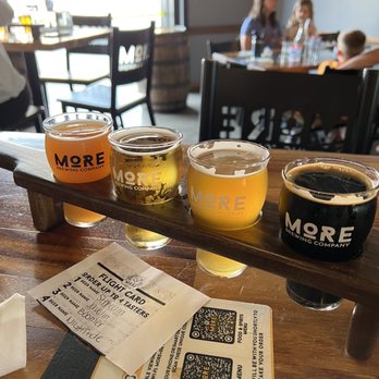 More Brewing - Four beers in different shades, from dark to light, are served in small glasses on a wooden table at More Brewing in Villa Park, IL. A flight card with handwritten notes and QR codes is placed nearby. The background shows a dining area with people, including children, seated at tables near large windows, with natural light illuminating the scene. - Brewpubs Near Me - Villa Park, Illinois
