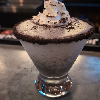 The Mill - Hershey - Cookies and Cream - A cookies and cream dessert presented in a clear glass, topped with a swirl of whipped cream and sprinkled with cookie crumbs. The dessert appears to have a creamy texture with dark cookie pieces mixed in. The background suggests a bar or restaurant setting, consistent with The Mill in Hershey, PA. - New American Near Me - Hershey, Pennsylvania