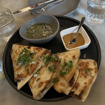 Root & Flower - Birria Quesadilla - A black round plate holds several slices of quesadillas topped with chopped green onions and cilantro. Two small bowls contain sauces, one with a brown sauce and the other with a green herb-based sauce. The plate is placed on a marble table, accompanied by glasses of water and utensils, suggesting a dining setting. - Wine Bars Near Me - Vail, Colorado