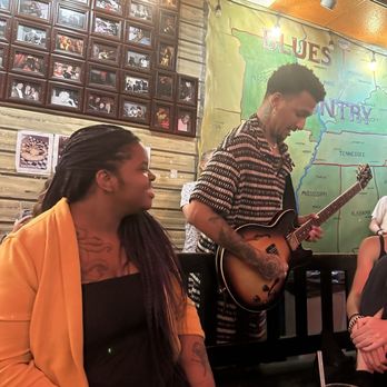 Kingston Mines - A man is playing an electric guitar in a venue decorated with a large map of the United States and a collage of framed photographs on the wall. A woman with long braided hair and a yellow jacket is seated nearby, smiling and looking at the musician. The setting appears to be a live music venue or bar with a casual atmosphere. - Jazz & Blues Near Me - Chicago, Illinois