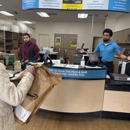 Spencer and ronni - Customer wearing a beige quilted jacket hands a brown Amazon package to an employee in a blue uniform at The UPS Store counter. The store interior includes shelving with supplies, a computer, and signage for services like printing and mailbox options. Two employees are visible behind the counter, one in a patterned shirt and the other in a blue shirt, with a curved overhead sign listing services.
