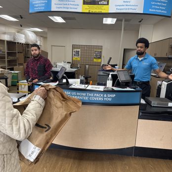 The UPS Store - Spencer and ronni - Customer wearing a beige quilted jacket hands a brown Amazon package to an employee in a blue uniform at The UPS Store counter. The store interior includes shelving with supplies, a computer, and signage for services like printing and mailbox options. Two employees are visible behind the counter, one in a patterned shirt and the other in a blue shirt, with a curved overhead sign listing services. - Printing Services Near Me - Olney, Maryland