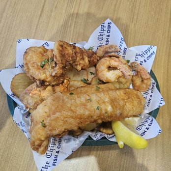 The Chippery - Union - A basket containing fried fish fillet, fried shrimp, and chicken wings, all coated in a crispy batter, is presented on branded paper from The Chippery - Union. The basket is lined with paper printed with the restaurant's name and description, and a lemon wedge is included as a garnish. The food is placed on a wooden table. - Fish & Chips Near Me - Union, New Jersey