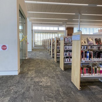 Central and Unser Library - Open and bright spaces - The image shows the interior of a library with bookshelves filled with books, a carpeted floor, and a ceiling with linear lighting fixtures. - Libraries Near Me - Albuquerque, New Mexico