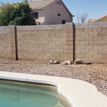 Jose Knows Trees - After 2 trees removed and stumps ground - The image shows a backyard with a swimming pool in the foreground, bordered by a concrete coping. Behind the pool, there is a gravel area with some rocks scattered along the edge. A tall brick wall encloses the yard, with residential houses visible beyond the wall. The scene is outdoors on a clear day with blue skies. - Tree Services Near Me - Chandler, Arizona