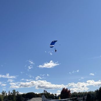 Skydive Oregon - A skydiver with a blue and white parachute is descending from the sky near a small airplane on the ground at an airfield. - Skydiving Near Me - Molalla, Oregon
