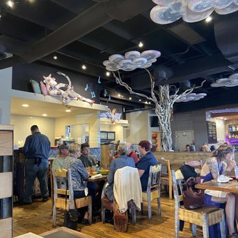 Range Cafe - Sunday at the Range Cafe! - The restaurant interior features a large artificial tree with twisted branches as a central decorative element. The ceiling is adorned with cloud-shaped art pieces, creating a whimsical atmosphere. Patrons are seated at wooden tables and chairs, engaging in conversation and dining. The background includes a visible kitchen area and a bar with bottles, with a dark ceiling and exposed ductwork. The overall setting is lively and creatively decorated. - Breakfast & Brunch Near Me - Albuquerque, New Mexico