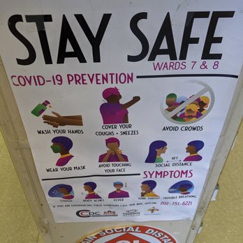 King's Cafe - COVID signage - A printed sign provides COVID-19 prevention guidelines and symptoms. It includes illustrations and text about washing hands, covering coughs and sneezes, wearing masks, avoiding touching the face, and maintaining six feet social distance. The sign also lists symptoms like cough, body aches, fever, sore throat, and trouble breathing. It features logos of health organizations and a hotline number. The sign is placed on a stand with a circular sticker below emphasizing social distancing of six feet. - Cafes Near Me - Washington, DC, District of Columbia