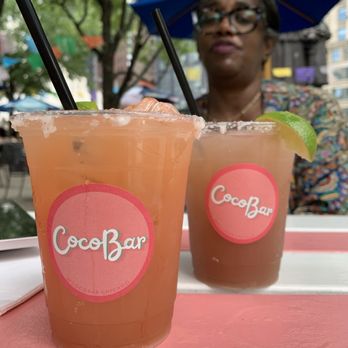 CocoBar - Two drinks with lime garnishes and black straws are placed on a pink and white table at CocoBar Chicago, with a woman in the background under a blue umbrella. - Latin American Near Me - Chicago, Illinois