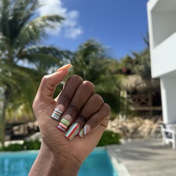Pick A Color Studio - line nail designs by Moet - A person's hand with multicolored striped nail designs is raised outdoors near a swimming pool, with palm trees and a building in the background. - Nail Salons Near Me - Brooklyn, New York