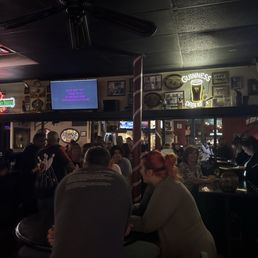 Thursday night Karaoke. - A lively bar scene with neon signs, a karaoke screen displaying song information, and patrons seated at tables and the bar, some singing or socializing.
