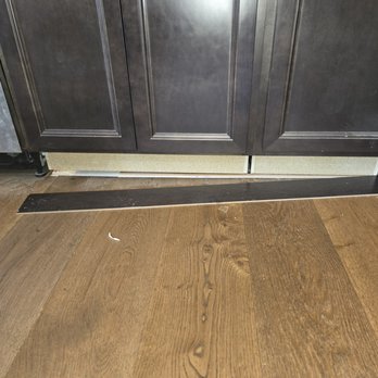 Three Rivers Builders - This is how they left my dishwasher in the hardwood floors just like that on top of it they damaged the dishwasher as well as our faucet. - The image depicts a section of a kitchen with dark-colored cabinets featuring raised panel doors. Below the cabinets, the baseboard is damaged or missing, exposing the wall or subfloor. The floor consists of wood planks with a natural finish, and there is a black strip or piece of trim on the floor near the base of the cabinets. Some small debris or paper fragments are visible on the floor. - Damage Restoration Near Me - Severna Park, Maryland
