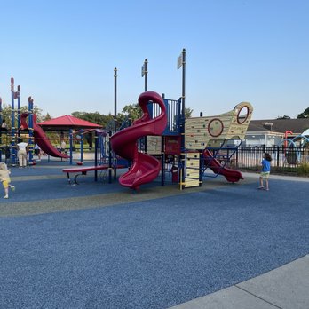 Prospect Park - The image depicts a playground at Prospect Park in Park Ridge, IL, featuring a large play structure with slides, climbing elements, and a ship-themed design. Children are playing on the equipment, and adults are present nearby. The playground surface is made of blue and green rubberized material, and the background includes a pavilion with a red roof, trees, and a clear blue sky. - Water Parks Near Me - Park Ridge, Illinois