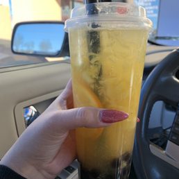 Hawaiian Fruit Tea with boba - A person holds a large plastic cup of yellow fruit tea with boba, inside a vehicle with a beige interior and side mirror visible.