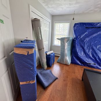 JK Moving Services - The image shows a room with various wrapped furniture pieces, boxes, and moving supplies. A large item is covered with a blue tarp, and other items are wrapped in protective material. There is a window allowing natural light, and a closet door is visible on the left side. The floor is wooden, and the ceiling has a textured finish. - Movers Near Me - Sterling, Virginia