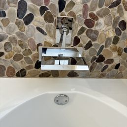 Installed waterfall faucet - A modern waterfall faucet is mounted on a pebble tile wall above a white bathtub, with a reflective chrome finish.