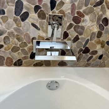 Eagle Design Build - Installed waterfall faucet - A modern waterfall faucet is mounted on a pebble tile wall above a white bathtub, with a reflective chrome finish. - General Contractors Near Me