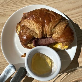 Apple-licious - Breakfast sandwich sausage - A breakfast sandwich featuring sausage, egg, and bacon on a croissant, accompanied by a small dish of honey, on a wooden table. - Bakeries Near Me - Spring, Texas