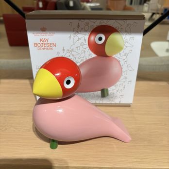 Design Warehouse - Decor - A pink bird-shaped figurine with a yellow beak and large eyes is placed on a wooden table, with its packaging box behind it. - Home Decor Near Me - Santa Fe, New Mexico