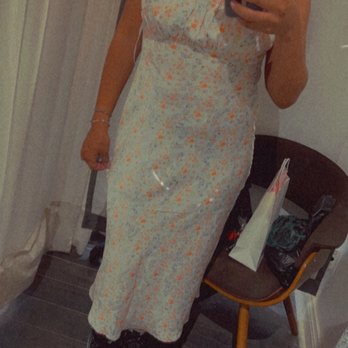 Pinkyotto - A woman with curly hair, wearing large hoop earrings, is taking a mirror selfie in a room. She is dressed in a sleeveless, floral-patterned dress and black sneakers. The room contains a chair with a bag and some items on it, and curtains are visible in the background. The image includes three fire emojis and a casual, stylish appearance. - Women's Clothing Near Me - Brooklyn, New York