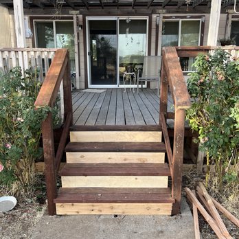 Outdoor Artisans - Back deck reinforcement with stairs replaced. - A wooden staircase with three steps, two of which are unfinished, leads up to a house porch. The porch has a sliding glass door and two windows, with outdoor furniture and decorations visible. Plants and bushes are on either side of the stairs, and the area appears to be in the process of renovation or construction. - Landscaping Near Me - Redding, California