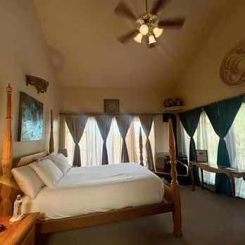 Black Bear Manor - Spacious room #10, second floor at the end of the hallway - The image shows a bedroom with a wooden four-poster bed with white bedding and pillows. There are curtains on the windows, some dark green and others light-colored, allowing natural light into the room. A ceiling fan with multiple lights is mounted on the ceiling. Additional furniture includes a nightstand with a lamp and water bottle, and a small table near the window. The room has a neutral color palette and decorative wall art. - Bed & Breakfast Near Me - Ouray, Colorado