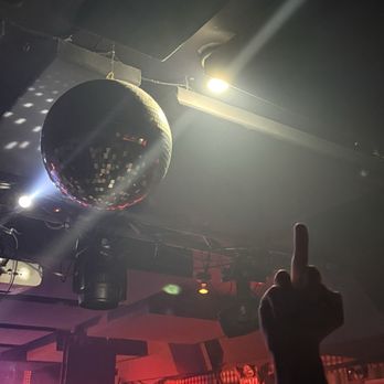 Baby's All Right - The image shows a person raising their middle finger in a nightclub setting. The background includes a large disco ball hanging from the ceiling, stage lighting, and a red-lit wall with an exit sign. The scene suggests a lively, nightlife environment with dim lighting and entertainment elements. - Bars Near Me - Brooklyn, New York