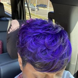 A person with short, curly purple hair is seated in the back seat of a vehicle, with their eyes closed and head tilted downward. The vehicle interior includes a headrest and a window showing a parking lot with several cars and a grassy area outside. The person is wearing a floral-patterned shirt, and sunlight is illuminating their hair and face.