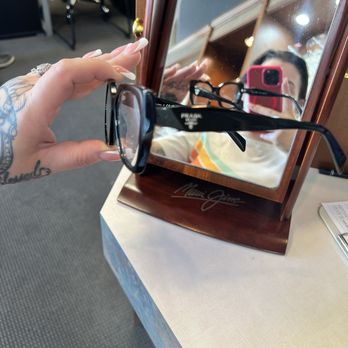 Lakeshore Optical - Went to the back for more glasses! - A person with tattoos holds Prada eyeglasses in front of a mirror, capturing their reflection with a smartphone. The mirror is on a wooden stand with a signature. - Eyewear & Opticians Near Me - Tempe, Arizona