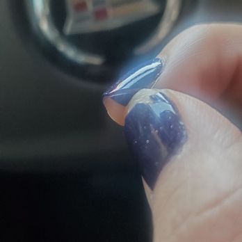 Super Nails - The image shows a person's hand with dark purple, glittery nail polish, positioned near the Cadillac emblem on a car's steering wheel. The focus is on the nails and the hand, with the Cadillac logo slightly blurred in the background. The setting appears to be inside a vehicle. - Nail Salons Near Me - Evergreen Park, Illinois