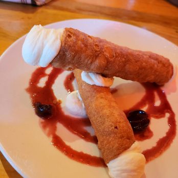 M'tucci's Italian - Cannoli - Two cannoli desserts are arranged on a white plate, with one cannoli positioned horizontally and the other leaning against it. The cannoli are topped with whipped cream, and the plate is decorated with berry sauce and whole berries. The setting appears to be a restaurant with a wooden table surface visible in the background. - Italian Near Me - Albuquerque, New Mexico