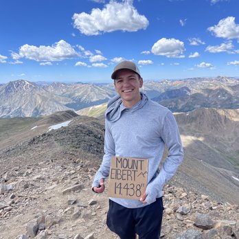 Mount Elbert - A smiling man in a gray hoodie and cap holds a sign indicating Mount Elbert's elevation of 14,438 feet, with mountain peaks and a blue sky behind him. - Hiking Near Me