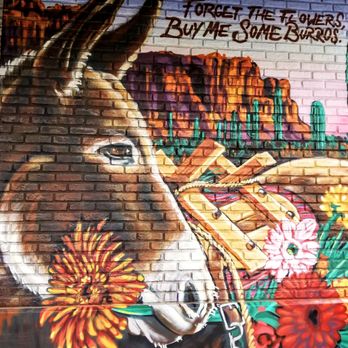 Someburros - The mural depicts a donkey's head with detailed shading and highlights, set against a desert landscape with mountains and cacti. The background includes graffiti-style text that reads, "Forget the flowers, buy me some burros." The artwork is painted on a brick wall, combining realistic animal portraiture with stylized desert scenery and urban graffiti elements. - Mexican Near Me - Mesa, Arizona