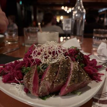 Prohibition - Fedora Fin (Cole slaw salad with Ahi tuna) - A plate of seared tuna with a seasoned crust is presented on a white dish, topped with shredded cheese and served alongside shredded vegetables. The setting appears to be a restaurant with a warm, dimly lit interior, and the table includes water glasses, a bottle, and utensils. The background shows other diners and ambient lighting. - Cocktail Bars Near Me - Murray, Utah