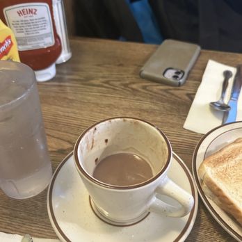 Salonica - Hot Chocolate - A ceramic cup with remnants of hot chocolate sits on a saucer on a wooden table, with a glass of water, toast, and condiments nearby. - Diners Near Me - Chicago, Illinois