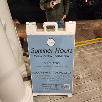 Lindy's Landing - A person stands outdoors at night near a sign for Lindy's Landing, showing summer hours and reservation info. - American Near Me - Wauconda, Illinois