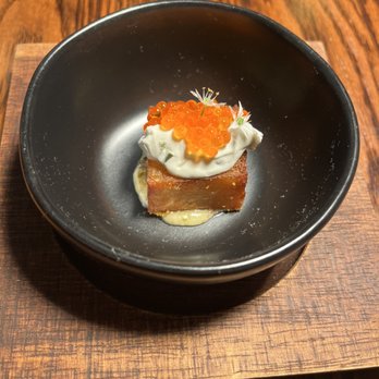 Michael's - Potato Pave, Toasted Seaweed Crema, Trout Roe - A gourmet appetizer presented in a black bowl on a wooden surface, consisting of a rectangular base, possibly a savory cake or terrine, topped with a dollop of cream, orange salmon roe, and small microgreens for garnish. The dish is arranged with attention to detail, highlighting contrasting textures and colors. - Seafood Near Me - St. Augustine, Florida