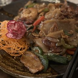 Drunken Noodle Lunch - A close-up of a plate featuring Pad See Ew, a Thai stir-fried noodle dish with wide rice noodles, vegetables such as carrots and green beans, and pieces of meat. The dish is garnished with a carved flower made from a vegetable and shredded carrots arranged decoratively on the side. Chopsticks are placed on the right side of the plate, and the background shows a blurred dining setting.