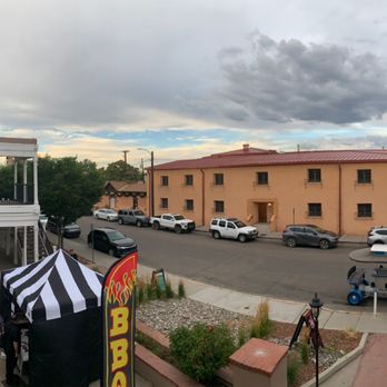 Downshift Brewing: Outpost 1706 - Outside view - A street scene featuring a two-story building with a balcony on the left, parked cars along the street, and a tent with a BBQ sign in the foreground. The sky is cloudy with some patches of blue, and there are people visible on the balcony and walking on the sidewalk. - Beer, Wine & Spirits Near Me - Albuquerque, New Mexico