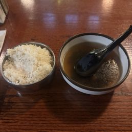 A bowl of clear, possibly Asian-style broth with a black spoon resting inside is placed on a wooden table. Next to it is a small metal cup filled with white rice. The setting appears to be in a restaurant or dining area with a warm, wooden surface.