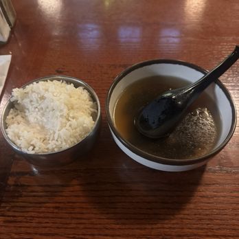 Yori - A bowl of clear, possibly Asian-style broth with a black spoon resting inside is placed on a wooden table. Next to it is a small metal cup filled with white rice. The setting appears to be in a restaurant or dining area with a warm, wooden surface. - Korean Near Me - Hurst, Texas