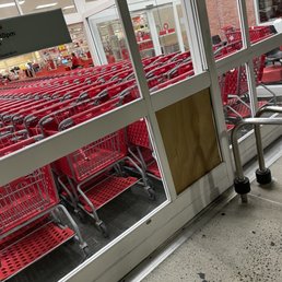 The window on the front door is broken. - The image shows a row of red shopping carts outside a Target store, separated by a glass door with a broken window panel. The carts are stacked and aligned, with some inside the store visible through the glass. The store interior features bright lighting and checkout counters. The broken window on the door is a notable feature, and the scene is captured from an angle that emphasizes the carts and the storefront.