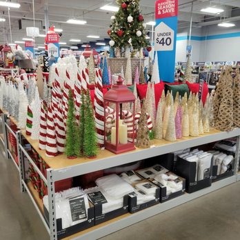 At Home - Small trees to add to your decor. - A variety of small Christmas trees, ornaments, and holiday decor are arranged on a display table in a store aisle, with signage indicating prices. - Home Decor Near Me - Albuquerque, New Mexico