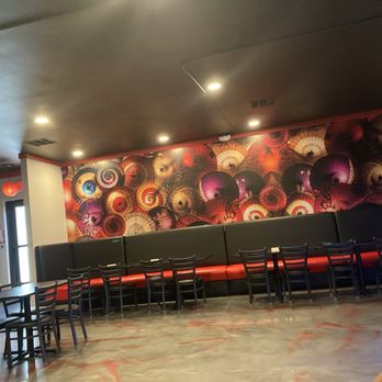 EZBachi - The image shows the interior of a restaurant with a mural wall decorated with numerous umbrellas in different colors and designs. There is a long black bench with red cushions along the mural wall, accompanied by several black chairs. The ceiling is dark, with recessed lighting, and the floor has a patterned surface. A door with a window and a small exit sign are visible on the left side of the image. - Teppanyaki Near Me - Phoenix, Arizona