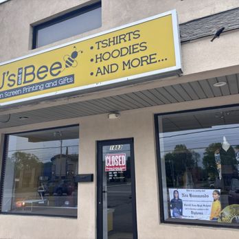 2 J's and a Bee - Front entrance. - The image shows the exterior of 2 J's and a Bee, a retail store specializing in custom screen printing and gifts. The storefront has a large yellow sign with black text listing products such as T-shirts, hoodies, and more. The shop's large glass windows display some items inside, and a door with a 'Closed' sign is visible. The building's facade is plain, with a small overhang above the entrance. The shop's address, 1883, is displayed on the door. - Screen Printing/T-Shirt Printing Near Me - Hermitage, Pennsylvania